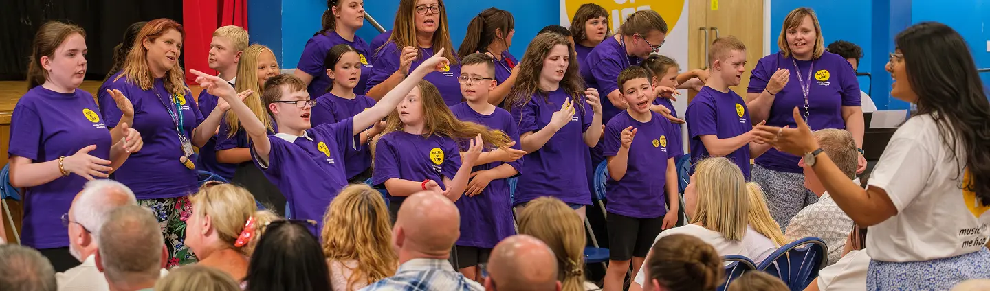 Group of children and adults wearing purple shirts perform sign language on stage at an event, facing an audience.