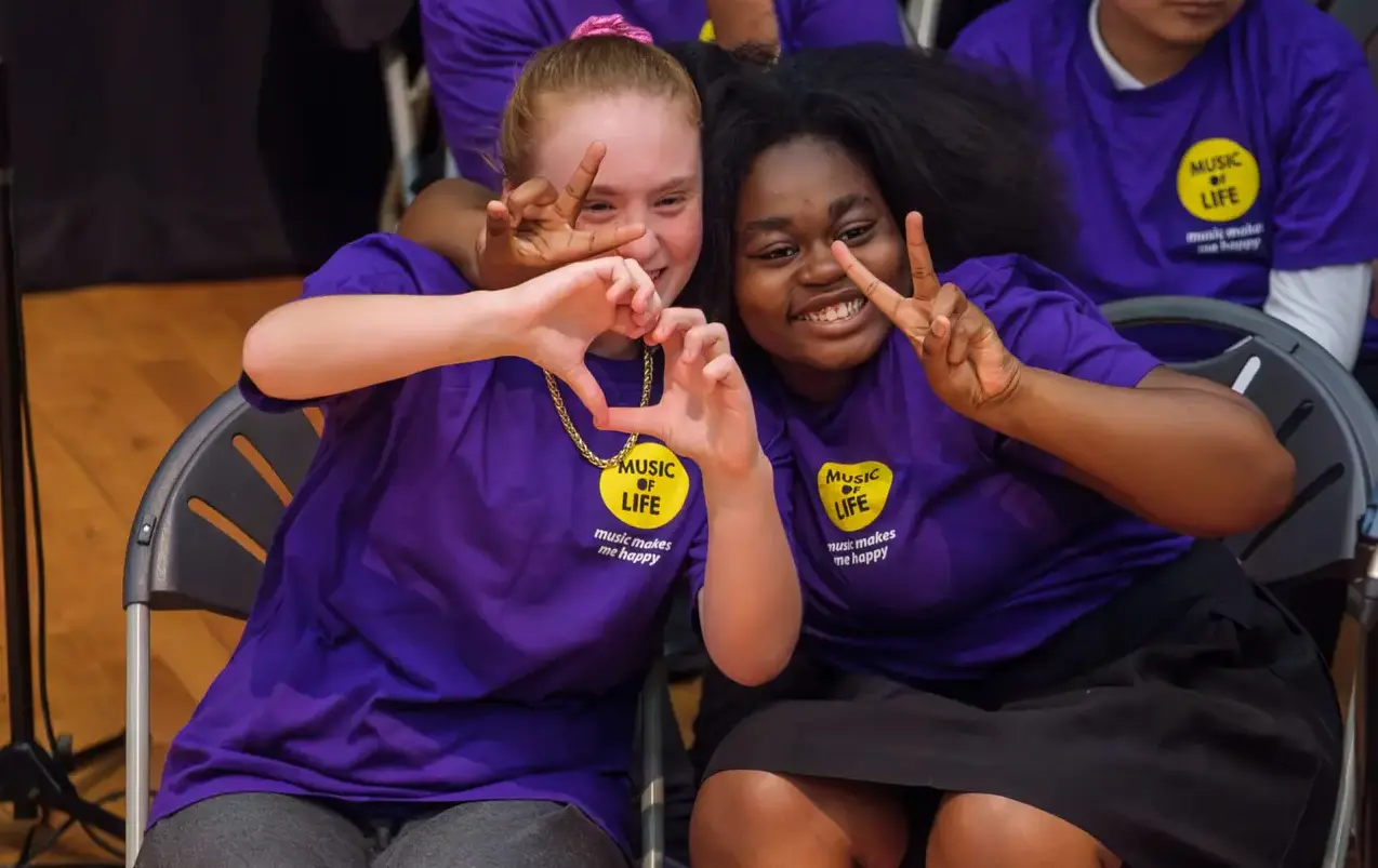 Two girls in purple shirts smiling and making peace signs at a music event, seated on metal chairs, wooden floor.