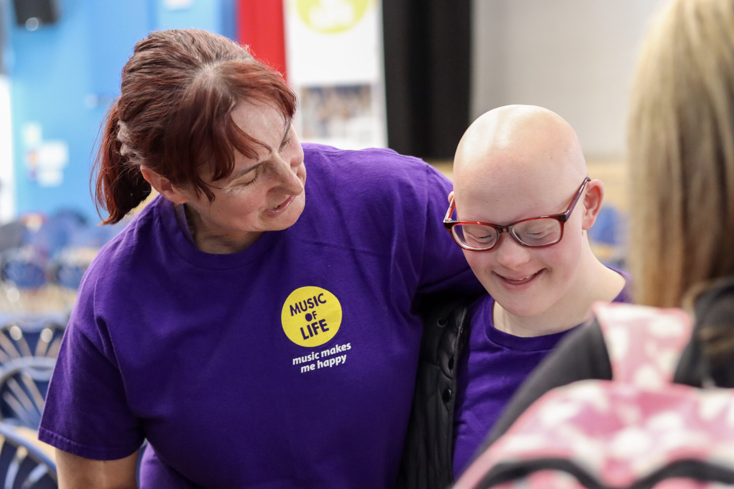A Music of Life music leader in a purple branded T-shirt smiles and leans towards a smiling young participant wearing glasses during a music session.
