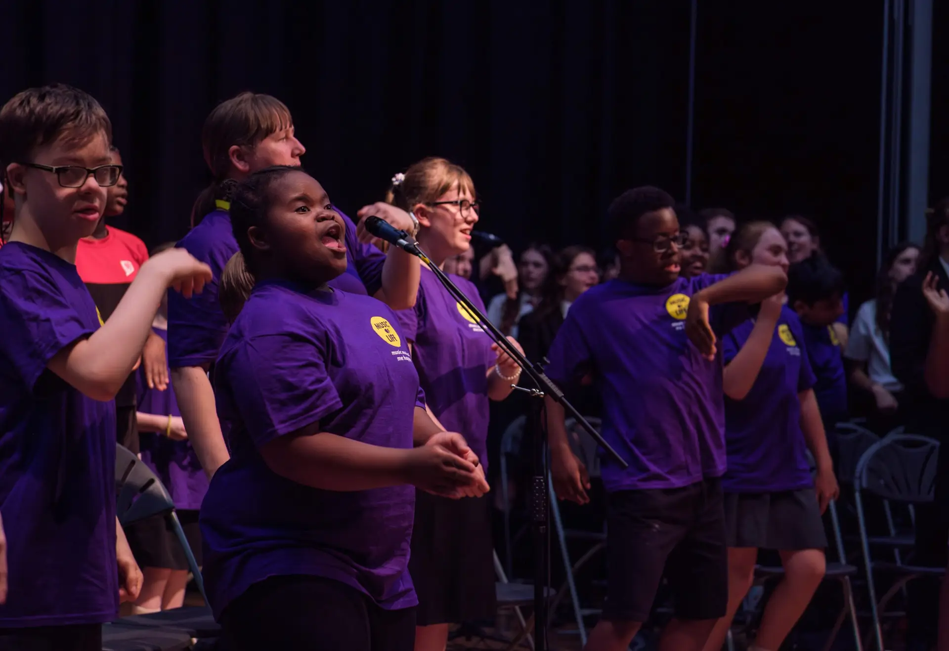 Children and young people in purple Music of Life T-shirts singing and signing together on stage at a concert.