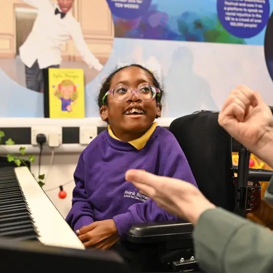 A smiling young girl wearing glasses and a purple school jumper sits in her wheelchair beside a keyboard during a music lesson. She looks towards an adult who is gesturing with their hands as they interact.